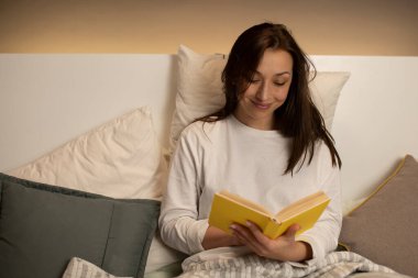 Güzel, siyah saçlı, pijamalı, kitap okuyan, sarı kapaklı, yatakta oturan bir kız.