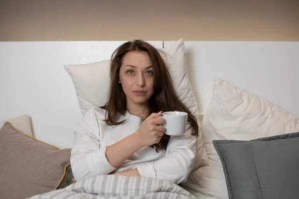 落ち着いてセクシーな女性がお茶を飲みながらベッドに座って、落ち着いてカメラを見て — ストック写真