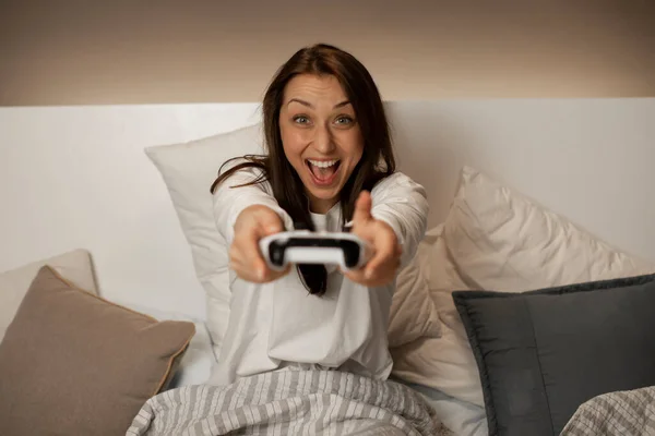 Joyeuse fille heureuse joue à des jeux vidéo avec joystick dans les mains tout en étant assis dans le lit — Photo