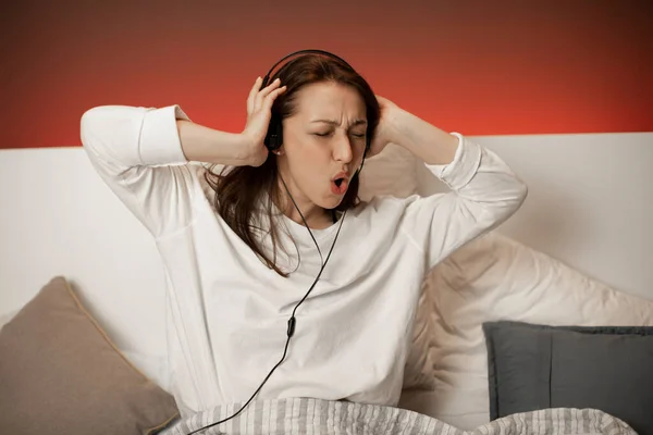 Linda chica de contenido en pijama escuchando música usando auriculares y bailando en la cama — Foto de Stock