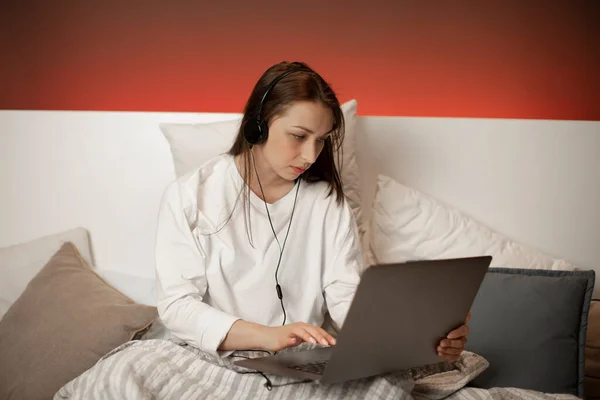 Pensativa chica de pelo oscuro en los auriculares sentados en la cama mirando su computadora portátil — Foto de Stock