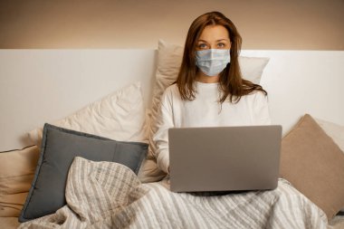 Yüz maskesi takmış çok çalışkan bir kız laptopta çalışıyor ve ne yazık ki kameraya bakıyor.