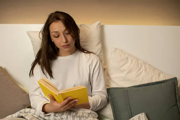 cute dark-haired girl in pajamas reading book with yellow cover sitting in bed