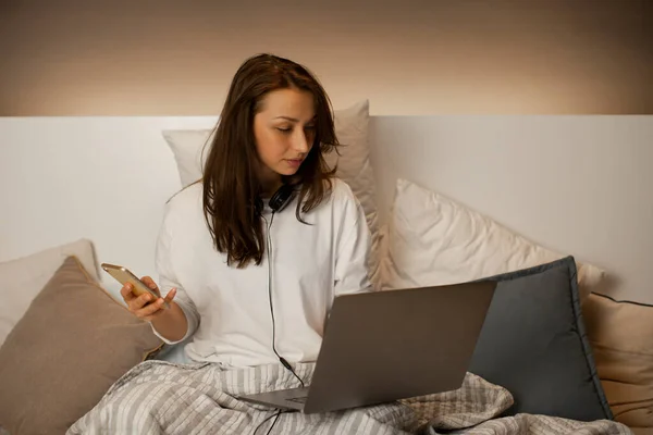 Atractiva chica de cabello oscuro sentada en la cama con su teléfono y su computadora portátil en un día perezoso — Foto de Stock