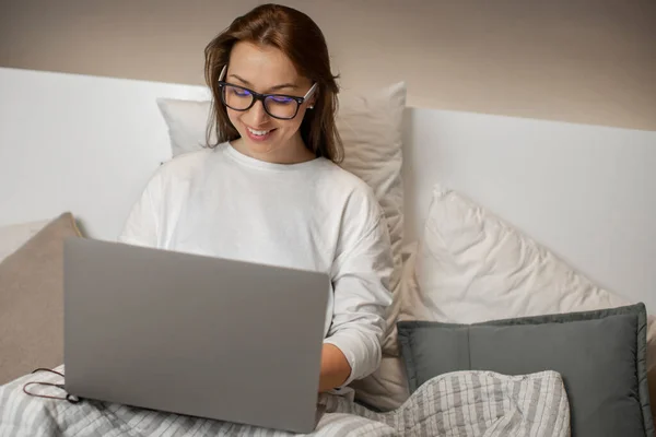 Bonita chica de pelo oscuro con gafas se sienta en la cama y sonríe mirando el ordenador portátil — Foto de Stock