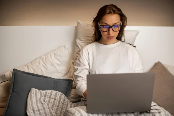 Mujer joven en gafas de trabajo en el ordenador portátil en la cama durante la cuarentena, freelance — Foto de Stock