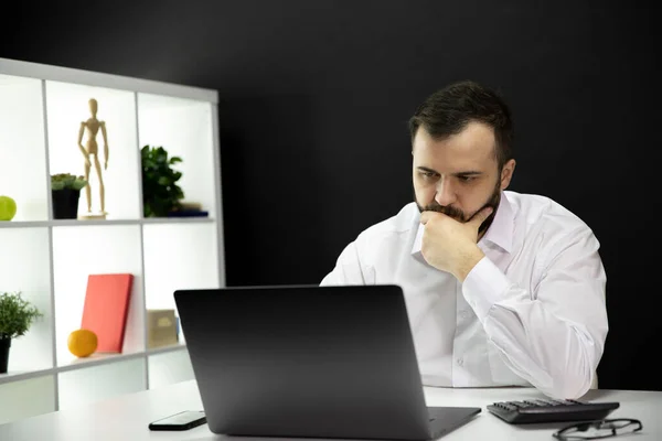 Freelancer desconcertado con la mano en la barbilla trabaja en el ordenador portátil en la oficina en casa, trabajo remoto — Foto de Stock