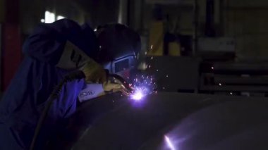 Koruyucu maskeli ve eldivenli kaynakçı endüstriyel bir atölyede büyük çaplı bir boruyu kaynaklıyor.