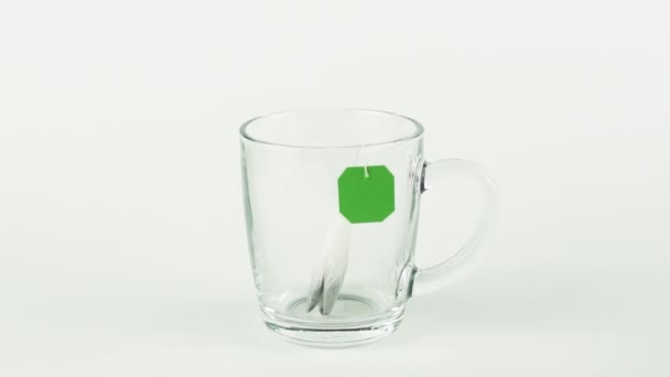 Black tea bag with green label is placed in glass transparent mug and filled with boiling water isolated on white background slow motion — Stock Video