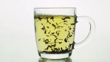 Siyah çay parçacıkları cam şeffaf bardağa dökülüyor ve beyaz arka planda izole edilmiş çay demlemek için kaynar suyla dolduruluyor.