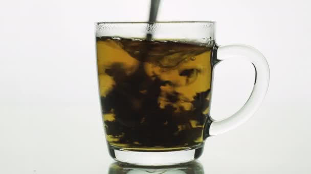 Kaşık, şeffaf cam bardakta çay karıştırır ve dönen siyah parçacıklar yaratır. Beyaz arkaplanda izole edildi — Stok video