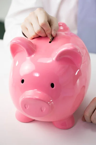 Cerca de mano masculina poner moneda a la alcancía rosa en el escritorio blanco, invertir concepto — Foto de Stock