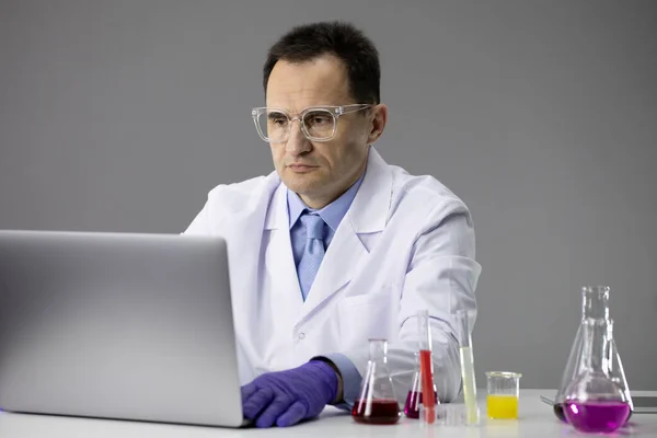 Kémiai kutató dolgozó lombikok töltött színes kémiai oldatok és laptop — Stock Fotó