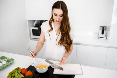 Genç, Güzel Kız Modern, Beyaz Mutfakta Kahvaltı Hazırlıyor. Omlet ve Baharatlı Sebzeler Tezgahta