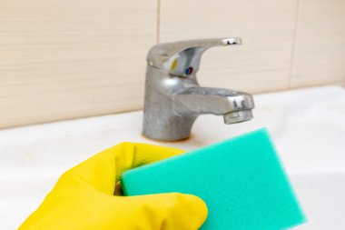Kirli musluğu kireç puluyla yıkamak için bir sünger tutan sarı lastik eldiven, limonlu su musluğu ve banyo lavabosunda limon tartısı olan ev temizleme konsepti.