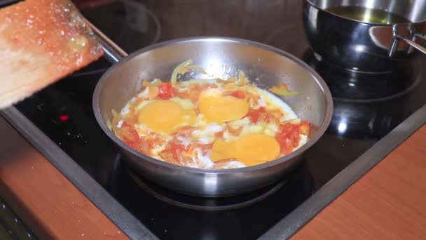 O chef cozinhar uma omelete, ovos mexidos em uma panela com um tomate picado e cebola, omelete de cozinha com legumes em casa de perto — Vídeo de Stock