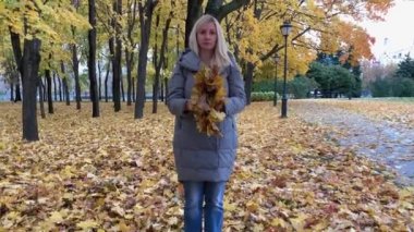 Sonbahar parkında duran ve bir yığın yaprak fırlatan kalın elbiseli sarışın genç bir kadın.