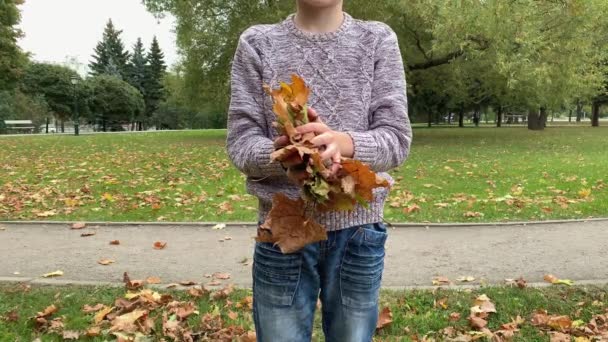 Неузнаваемый подросток бросает сухие кленовые листья в осенний парк — стоковое видео