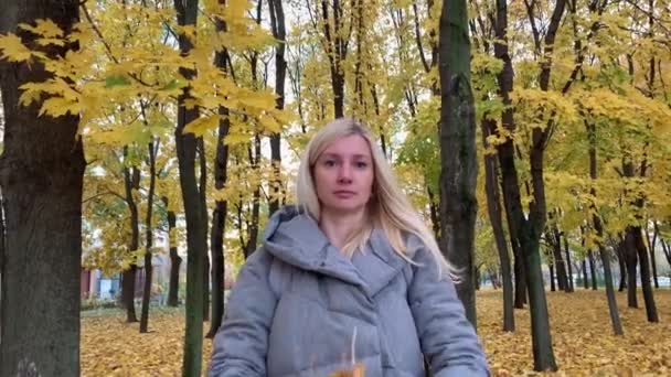 Młoda blondynka w ciepłych ciuchach stojąca jesienią w jesiennym parku i rzucająca kupą liści klonu — Wideo stockowe