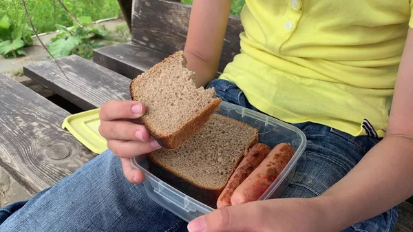 Preteen chlapec jíst kus celozrnného hnědého chleba z plastu vyndat kontejner s grilovanými klobásami, zatímco sedí na dřevěné šicí pilky houpačka na otevřeném vzduchu, venkov piknik koncept — Stock fotografie