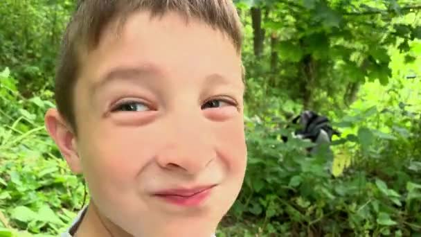 Un retrato de un niño preadolescente sonriendo en cámara de cerca, se sienta en el suelo en el bosque de verano y el follaje verde en el fondo — Vídeos de Stock