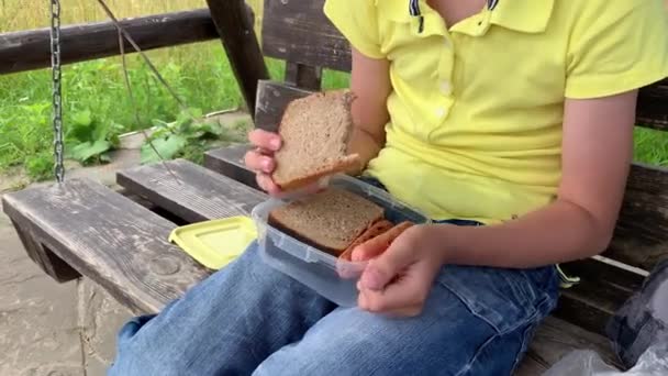 En ung pojke äter en bit fullkornsbrunt bröd från en plastburk med grillad korv medan han sitter på en träsåg svingande säng på det fria, lantliga picknickkonceptet — Stockvideo