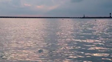 Yaz günbatımında sakin deniz suyu, ufukta küçük dalgalar, iskele ve deniz feneri olan su yüzeyinde güneş yansıması.