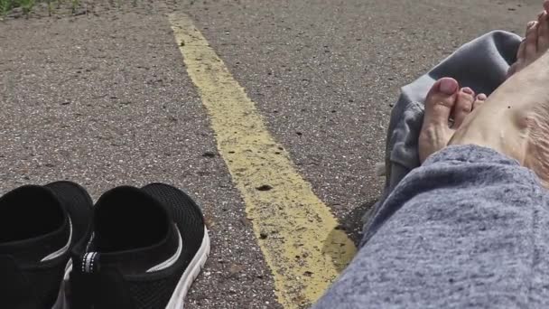 Woman with bare feet and backpack sitting on an asphalt road with a yellow single road line waiting vehicle for hitchhiking, hiking, summer season — Stock Video