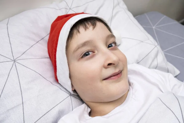 Egy fiatal fiú fekszik az ágyban télapó kalapban karácsony este, és várja az ajándékokat és csodákat, boldog karácsonyt és újévi ünnepet — Stock Fotó