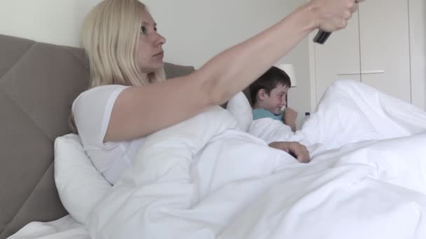 Eine blonde Frau liegt im Bett und schaltet die Kanäle per Fernbedienung, ihr Sohn im Hintergrund spielt Spiele auf dem Smartphone — Stockvideo