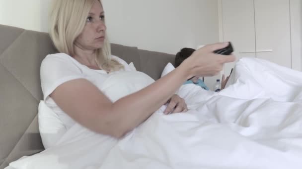 Блондинка, лежащая в постели и переключающая каналы с помощью дистанционного управления, ее сын на заднем плане играет в игры на смартфоне — стоковое видео