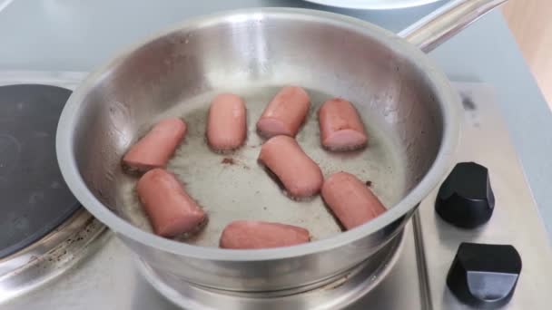 Een womans hand frituren worsten in grote hoeveelheid olie in een stalen pan op een elektrische kachel, en draaien het met een spatel voor het ontbijt thuis — Stockvideo