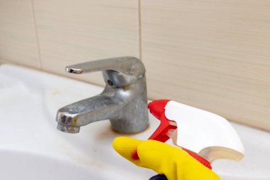 Kirli musluğu kireç puluyla yıkamak için sprey temizleme maddesi tutan sarı lastik eldivenler, limonlu su musluğu, banyo lavabosu, ev temizliği konsepti.