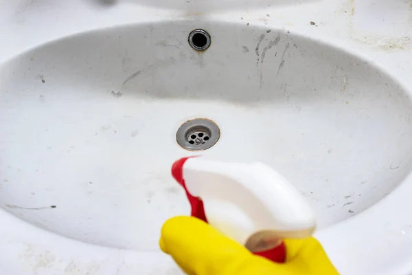 Рука в резиновой желтой перчатке с помощью распылителя для очистки и дезинфекции грязной ржавой посуды, раковины с лимфорамой и мыльными пятнами — стоковое фото