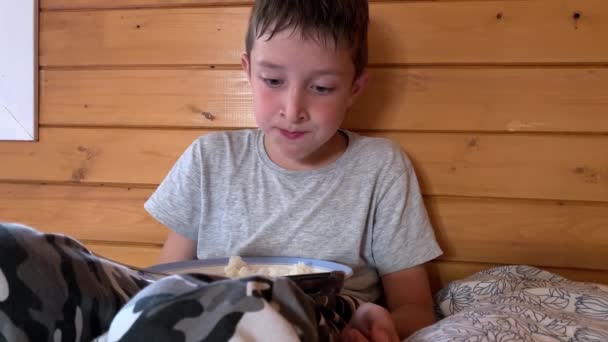 Um menino faminto e faminto com o cabelo molhado sentado em uma cama em uma casa de madeira rural e comendo arroz gananciosamente, ele jogou fora e ficou com muita fome. — Vídeo de Stock