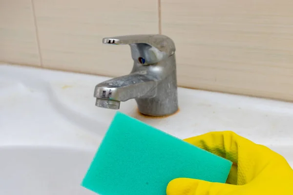 Hand in yellow rubber gloves holding a sponge for washing dirty faucet with limescale, calcified water tap with lime scale on washbowl in bathroom, house cleaning concept.