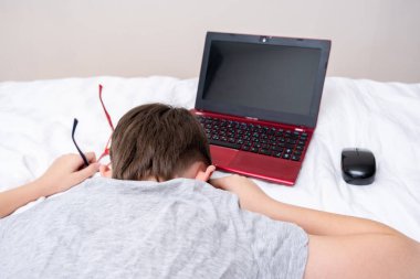 Sıkıcı ve yorgun çocuk ödevini yaptıktan sonra dizüstü bilgisayarla yatakta uzanıyor. Karantina sırasında uzaktan eğitim alıyor..