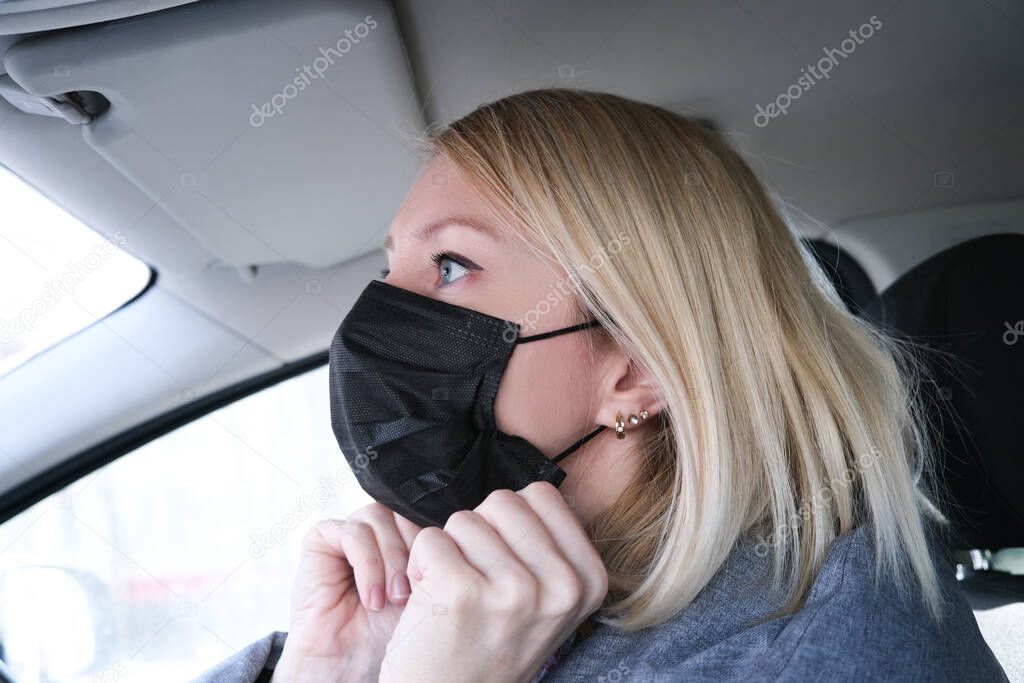 Caucasian european woman sitting in a car and putting on black surgical medical face mask as a way of protection against coronavirus.
