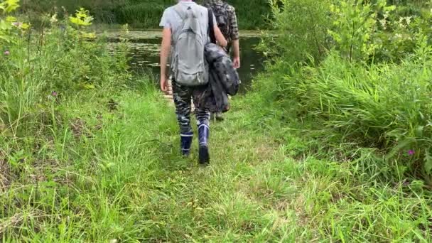 Mãe e filho turistas com mochilas, caminhantes caminhando para o rio ao longo do caminho coberto com grama alta e admirando a natureza e água calma no verão — Vídeo de Stock