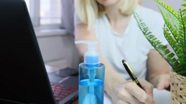 Bulanık yüzlü kadın maske takıyor. Evde dizüstü bilgisayarda çalışıyor. Koruyucu olarak el dezenfektanı var.