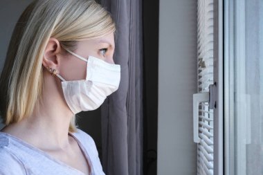 Cerrahi maske takmış korkmuş bir kadın evde kalıp pencereden dışarı bakıyor. Karantina ve kendini izole etme, koronavirüs patlaması ve epidemi..
