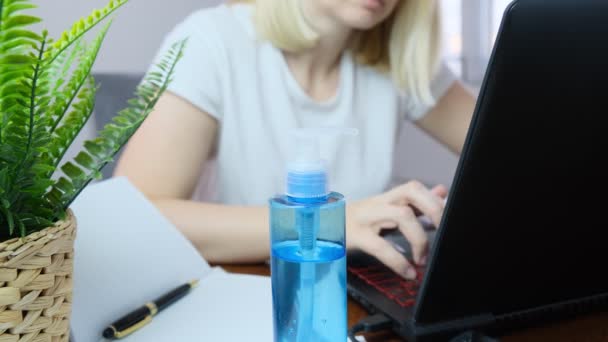 Mujer borrosa freelancer en la cara máscara médica que trabajaen un ordenador portátil en casa, desinfectante de manos como una protección está sobre la mesa delante de ella — Vídeos de Stock