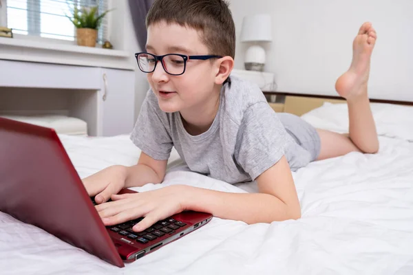 A boy, school boy lying in bed with a laptop and watching online translation, lesson, e-learning and education concept.