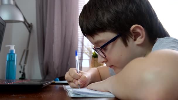 Um menino, estudante sentado à mesa, mesa com laptop e fazendo lição de casa em casa, e-learning, lição distante e conceito de educação on-line — Vídeo de Stock