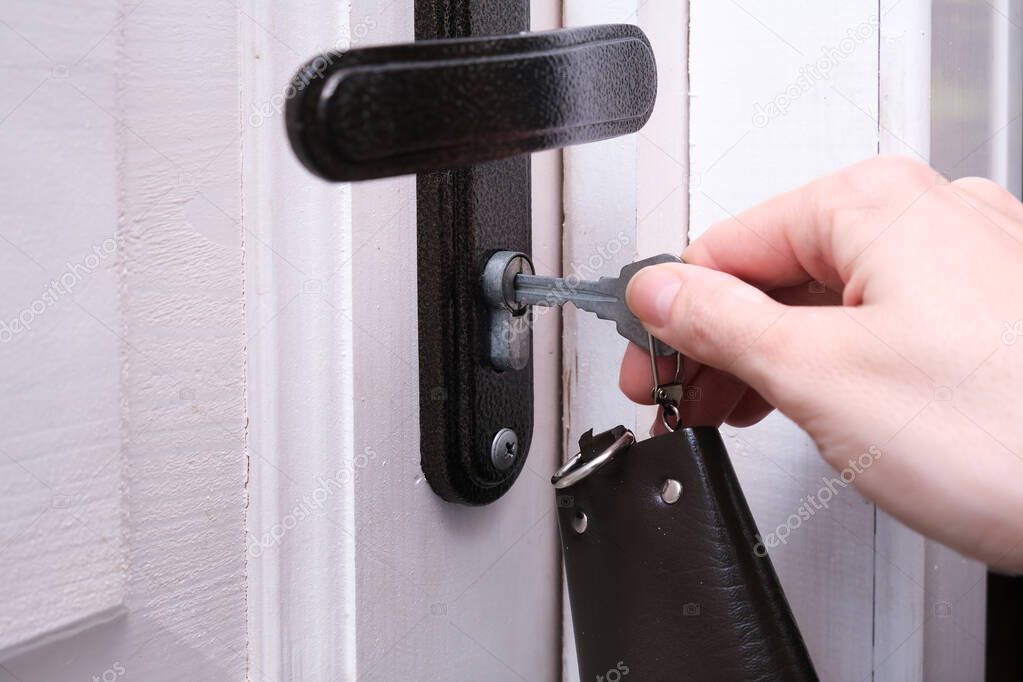 A hand holding a key and inserting it in a keyhole in order to open or lock a white wooden door, home security and protection concept.