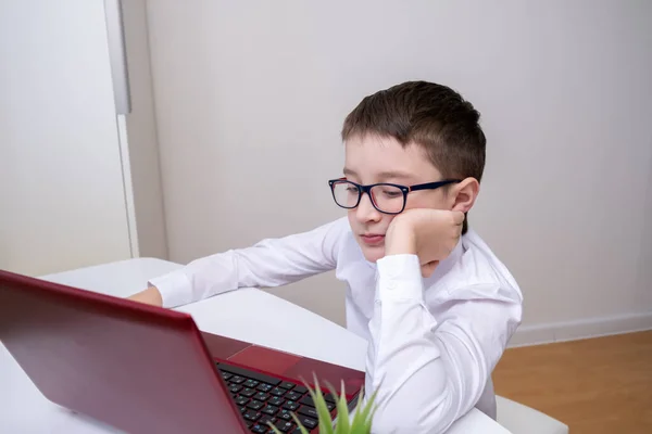 A boy, schoolboy in white formal shirt sitting by the table, desk with laptop and doing homework at home, e-learning, distant lesson and online education concept.