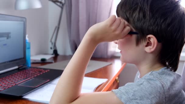 Un niño, colegial sentado junto a la mesa, escritorio con portátil y haciendo deberes en casa, e-learning, lección a distancia y concepto de educación en línea — Vídeo de stock