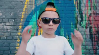 Bir kap ve graffiti ile duvarın yanında kamera poz güneş gözlüğü serin 5 yaşında bir çocuk, bir çocuk güneş gözlüğü onları ve onları geri giysiler alır