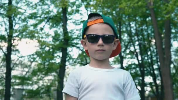 Een coole jongen in zonnebril toont een klasse met zijn vinger omhoog staande in een park van zomer tegen een achtergrond van groene bomen — Stockvideo