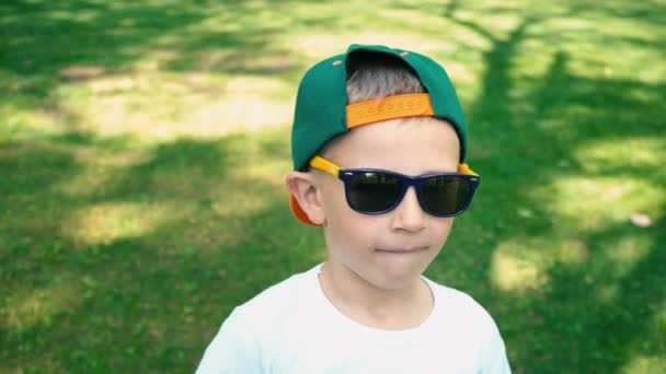 Un garçon frais de 5 ans en lunettes de soleil se tient dans le parc d'été et commence à rire de quelqu'un, les vraies émotions de l'enfant — Video